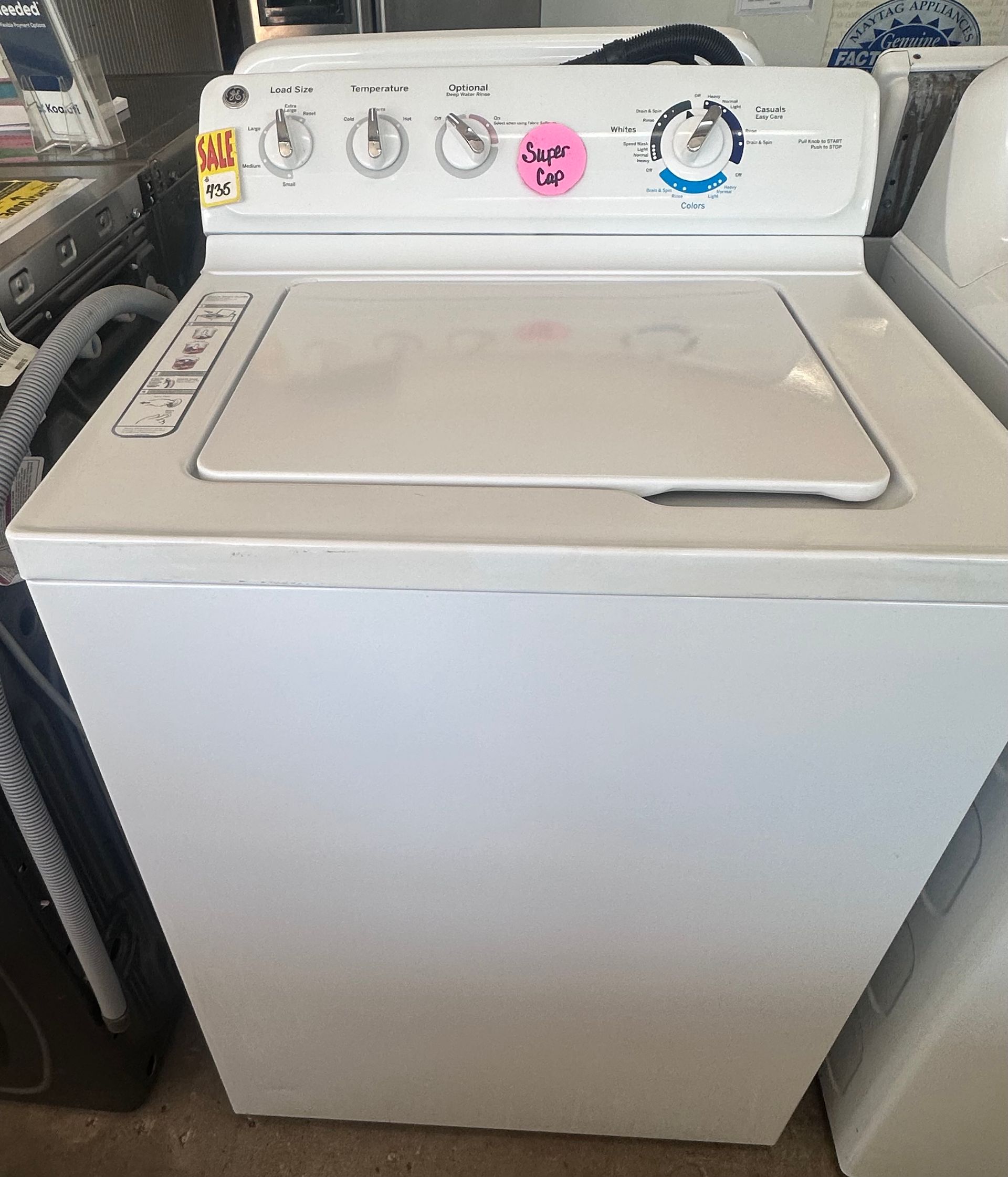 White top-load washing machine in a laundry room, with control knobs and a pink sticker on the panel.