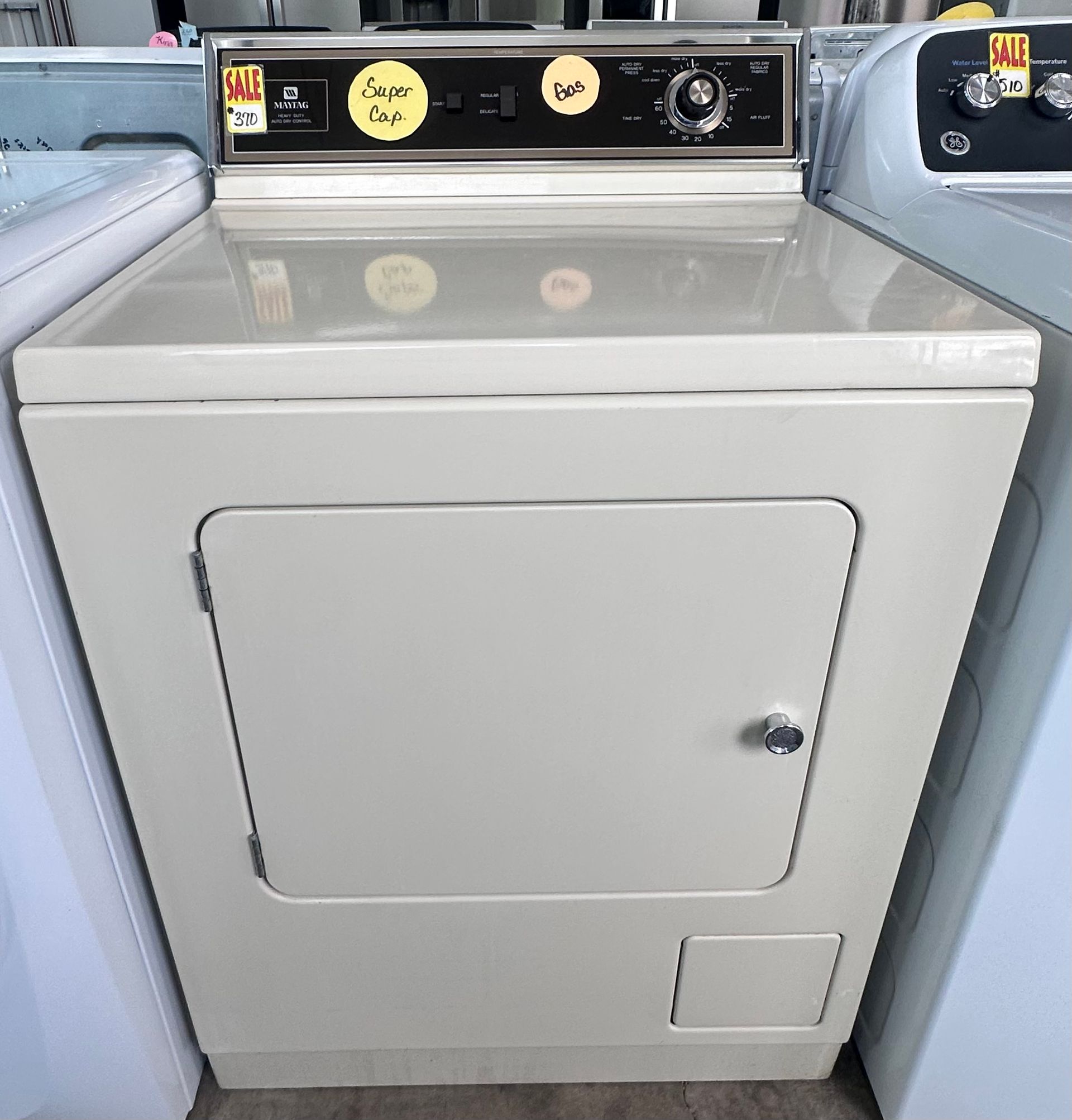 A white top-loading washing machine with a control panel featuring five knobs, displayed in a retail showroom.