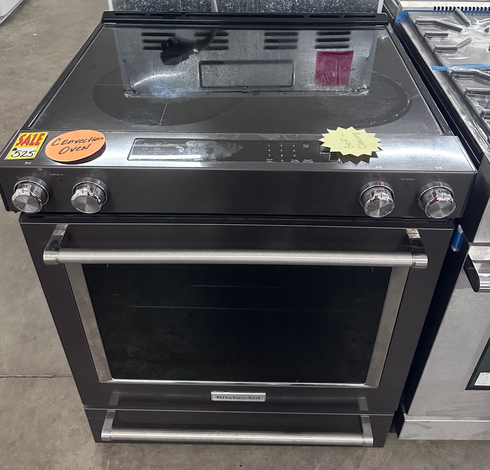 Black stainless steel oven with glass door and four front knobs on a store floor