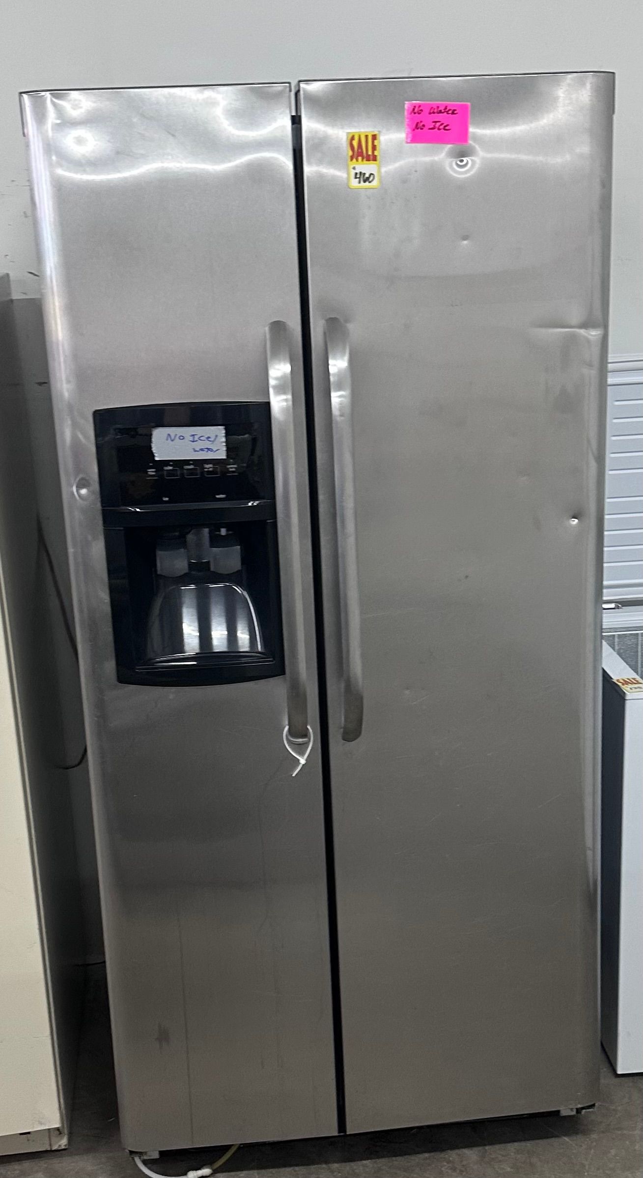 Stainless steel side-by-side refrigerator with water and ice dispenser, shown in a store aisle.