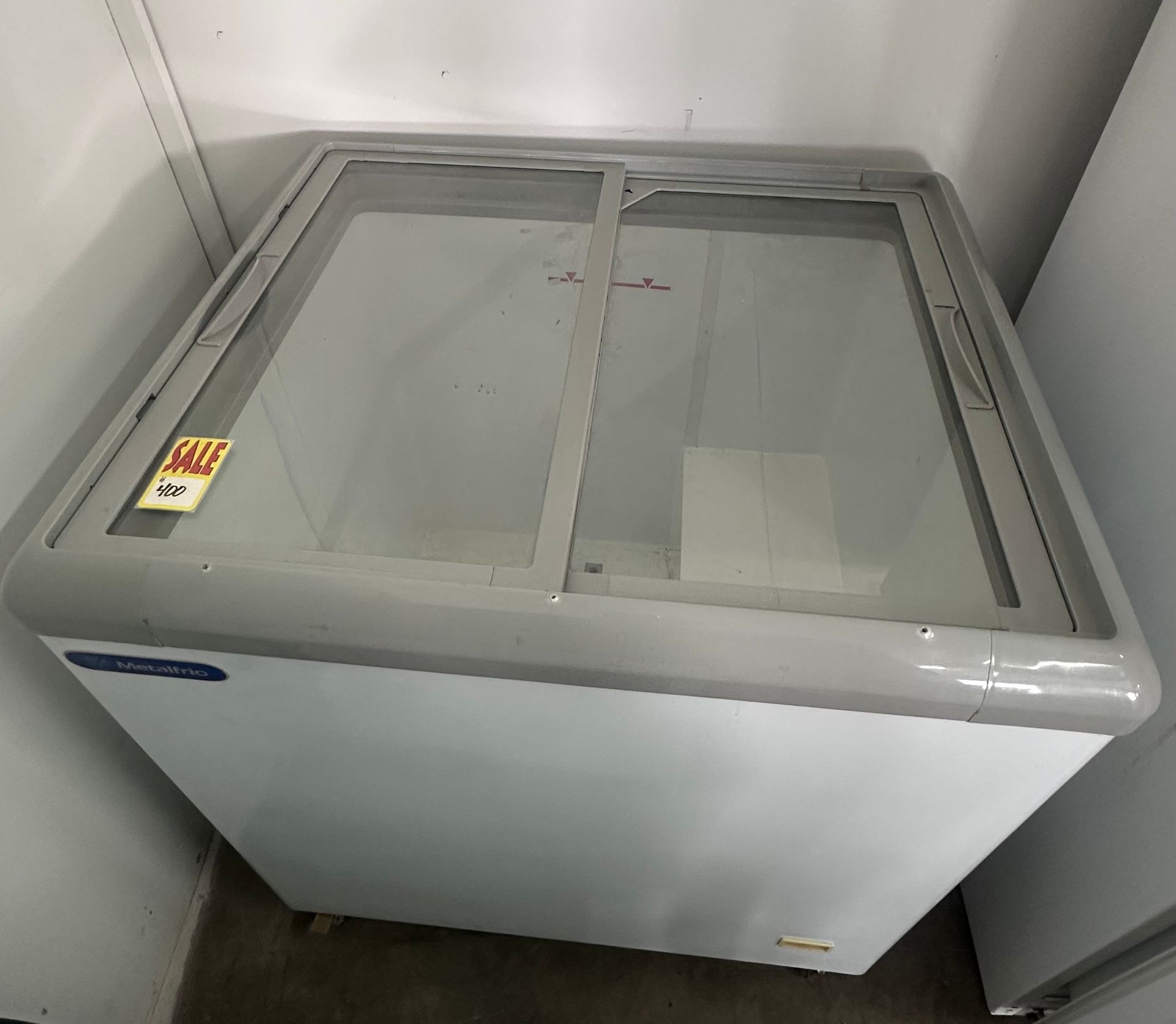 White chest freezer with sliding glass lids in a corner against plain walls.