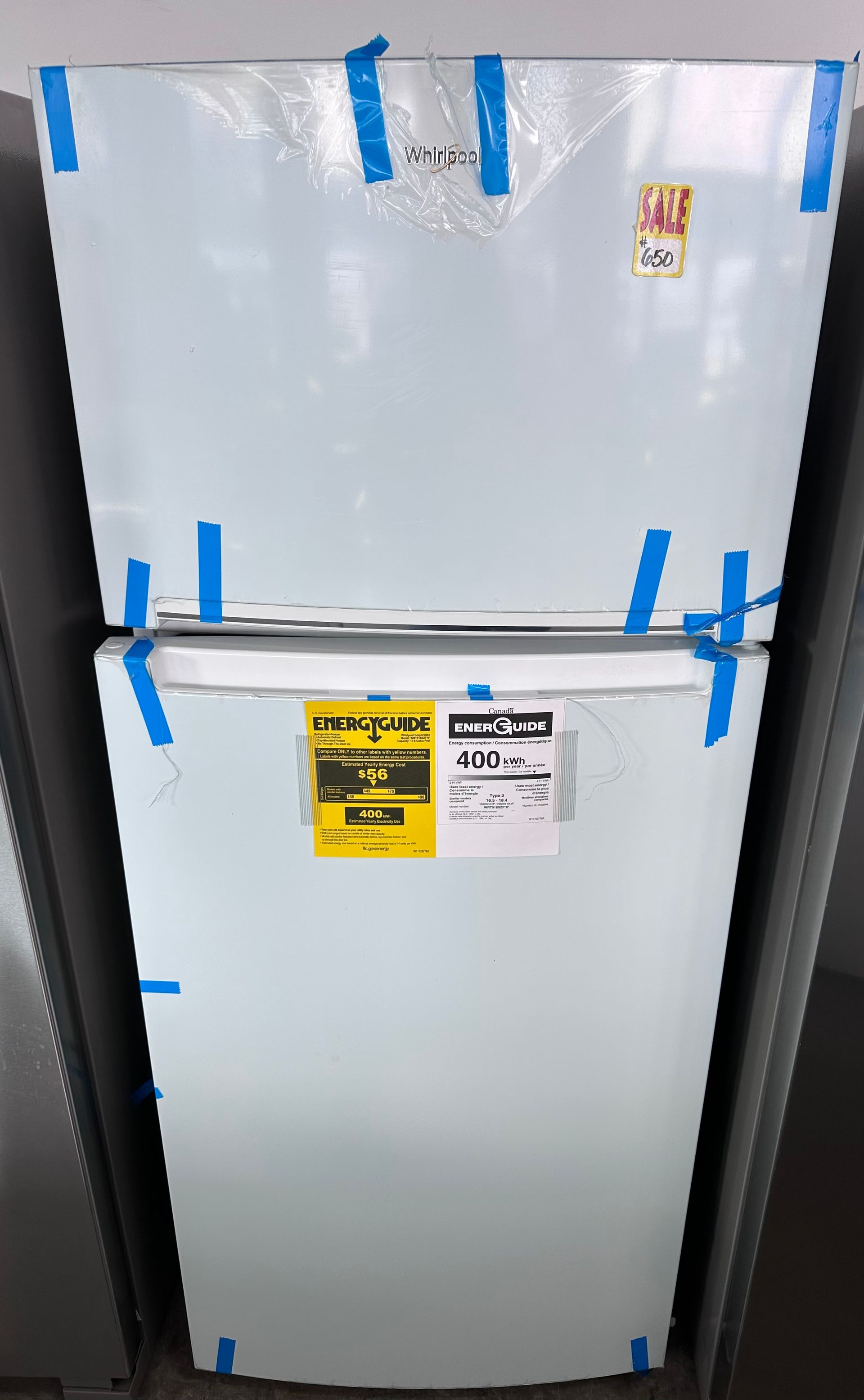 White refrigerator, top-freezer style, with blue tape securing doors. A yellow energy efficiency sticker and sale tag are visible.