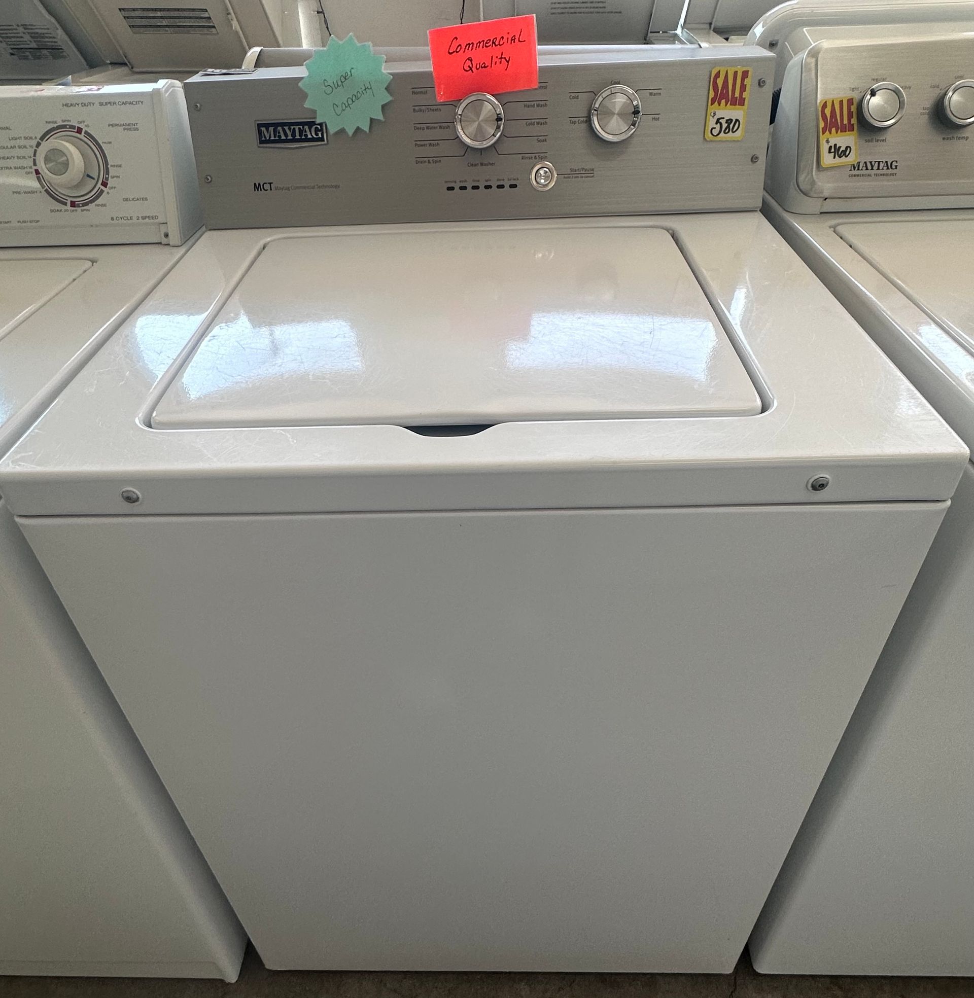 White top-load washer with a front control panel in a laundry store aisle; dryers are visible on both sides.
