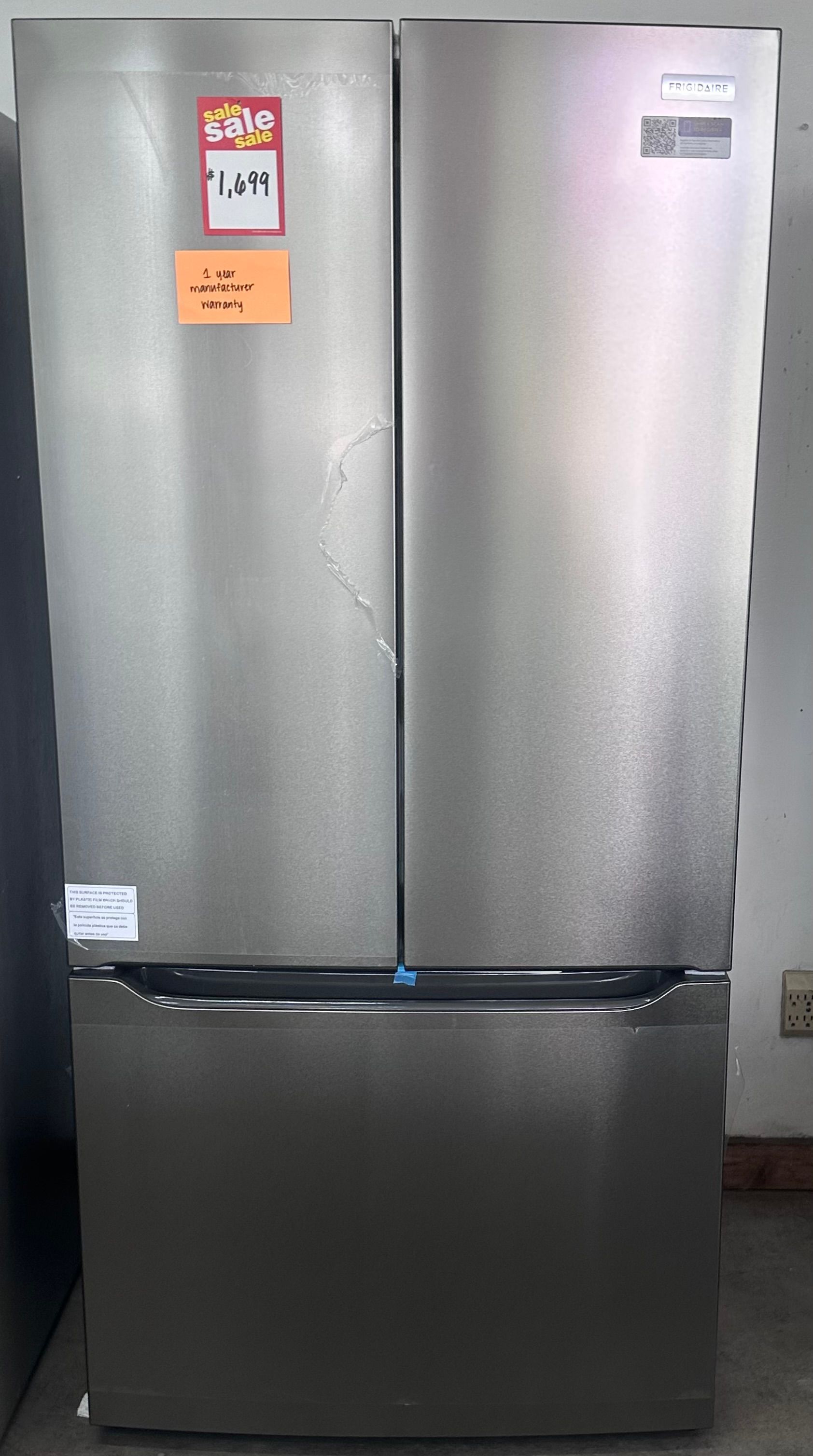 Silver double-door refrigerator standing against a dark wall with a black freezer drawer at the bottom