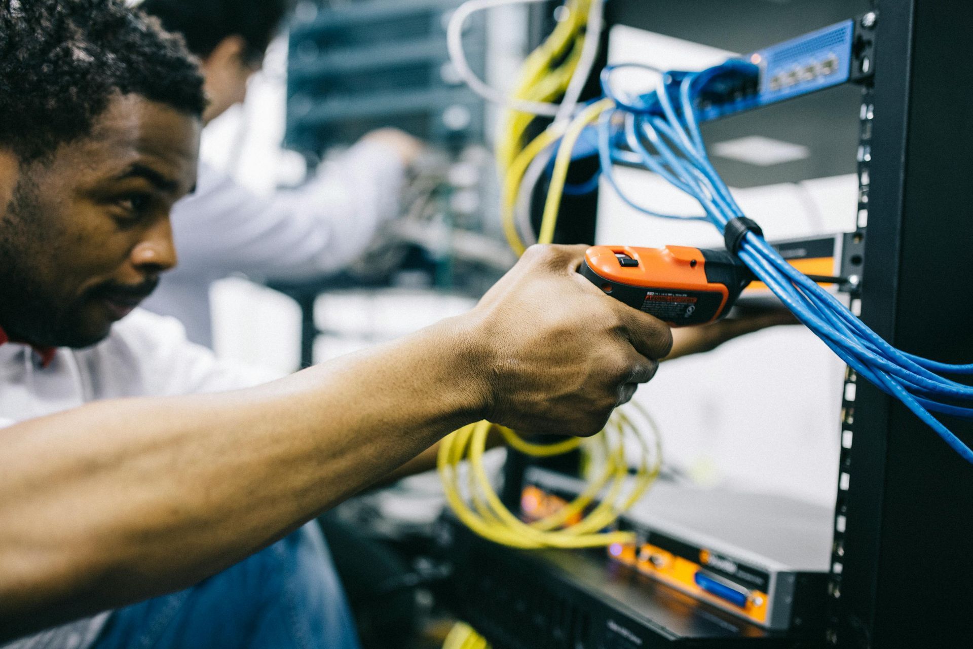 Man working on server cables with an orange tool in a server room, with another person in the background.