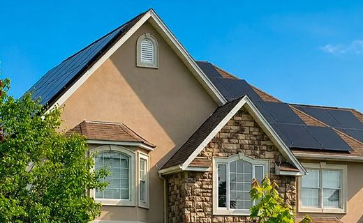 Suburban house with beige siding, stone accents, and solar panels on the roof under a blue sky