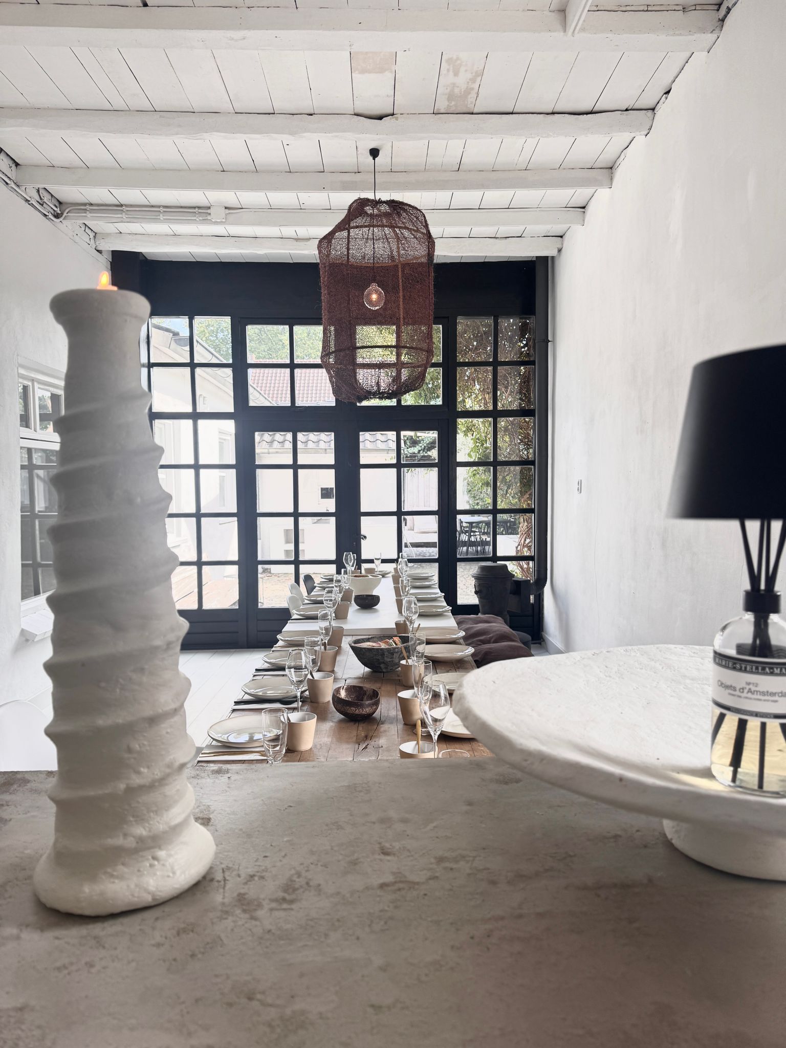 Een lange eettafel gedekt voor een maaltijd, in een witgekalkte kamer met een kroonluchter.