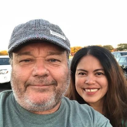 A man wearing a grey patterned baseball cap and a woman with long dark hair smiling together outdoors.