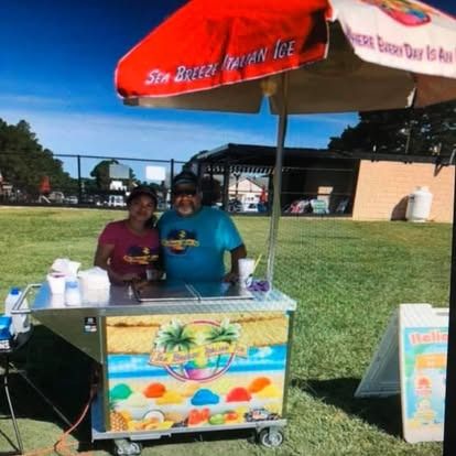 A man and a woman stand smiling behind a Sea Breeze Italian Ice cart with a red umbrella on a grassy field.