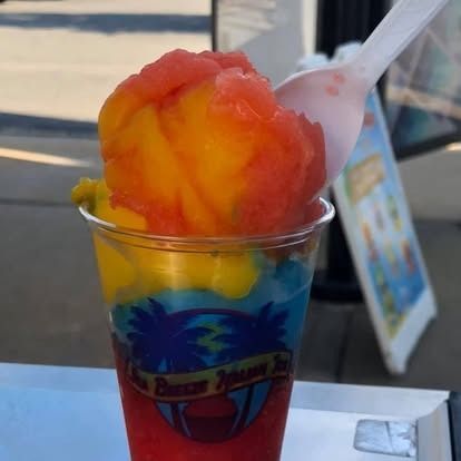 A plastic cup filled with colorful slushy layers of red, yellow, and blue, topped with a spoon.