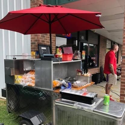 A portable metal food cart with a red umbrella sits on a sidewalk outside a brick building, with a person standing nearby.