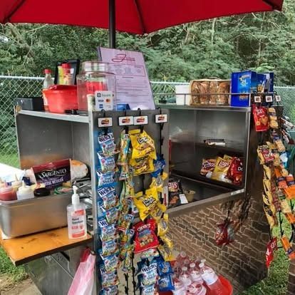 A food cart with a red umbrella, displaying a menu, various snacks, and drink supplies for sale outdoors.