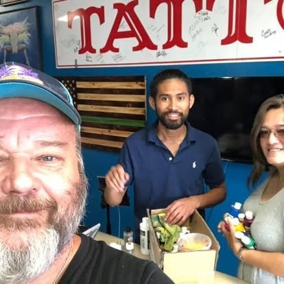 Three people pose together inside a tattoo shop, standing near a counter with various supplies.