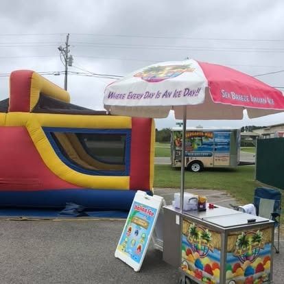 A colorful bounce house next to an ice cream stand under a branded umbrella in a parking lot.