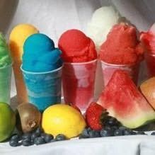 Colorful scoops of shaved ice or sorbet in plastic cups, arranged with fresh fruit like watermelon, strawberries, and limes.
