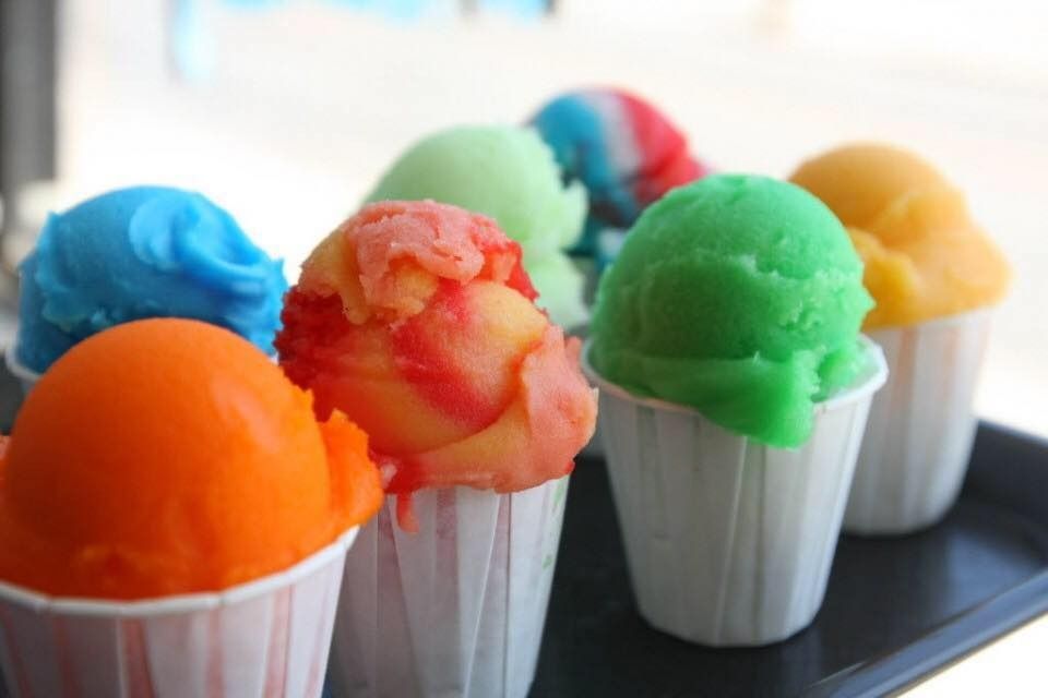 Colorful scoops of fruit sorbet or ice cream served in small white paper cups on a black tray.