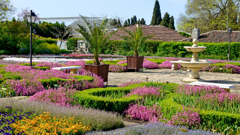 Formal garden with pink flowers, brick paths, potted palms, and a tiered fountain under a bright blue sky