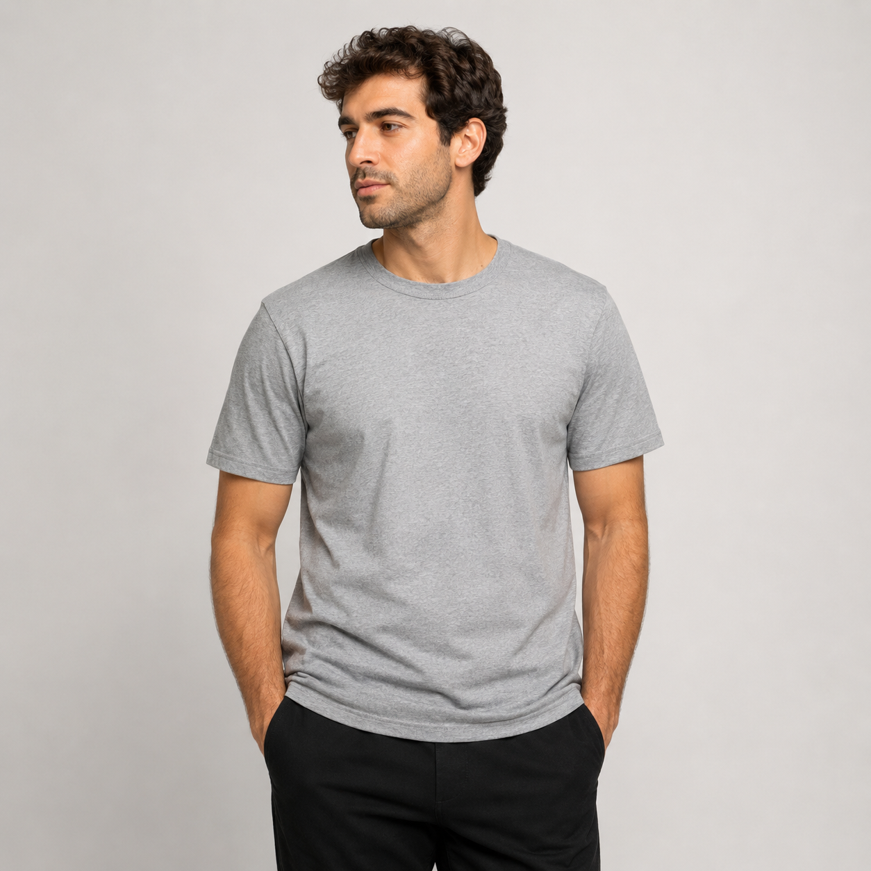 Hombre con camiseta gris y pantalón negro, de pie con las manos en los bolsillos, mirando hacia un lado.
