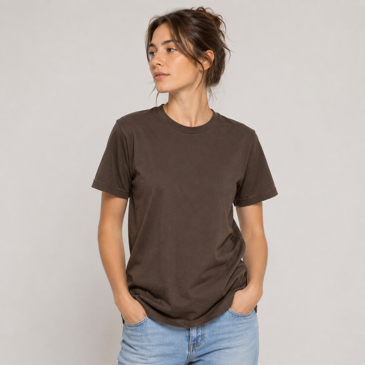 Mujer con camiseta marrón oscuro y vaqueros, de pie sobre un fondo liso, con las manos en los bolsillos.