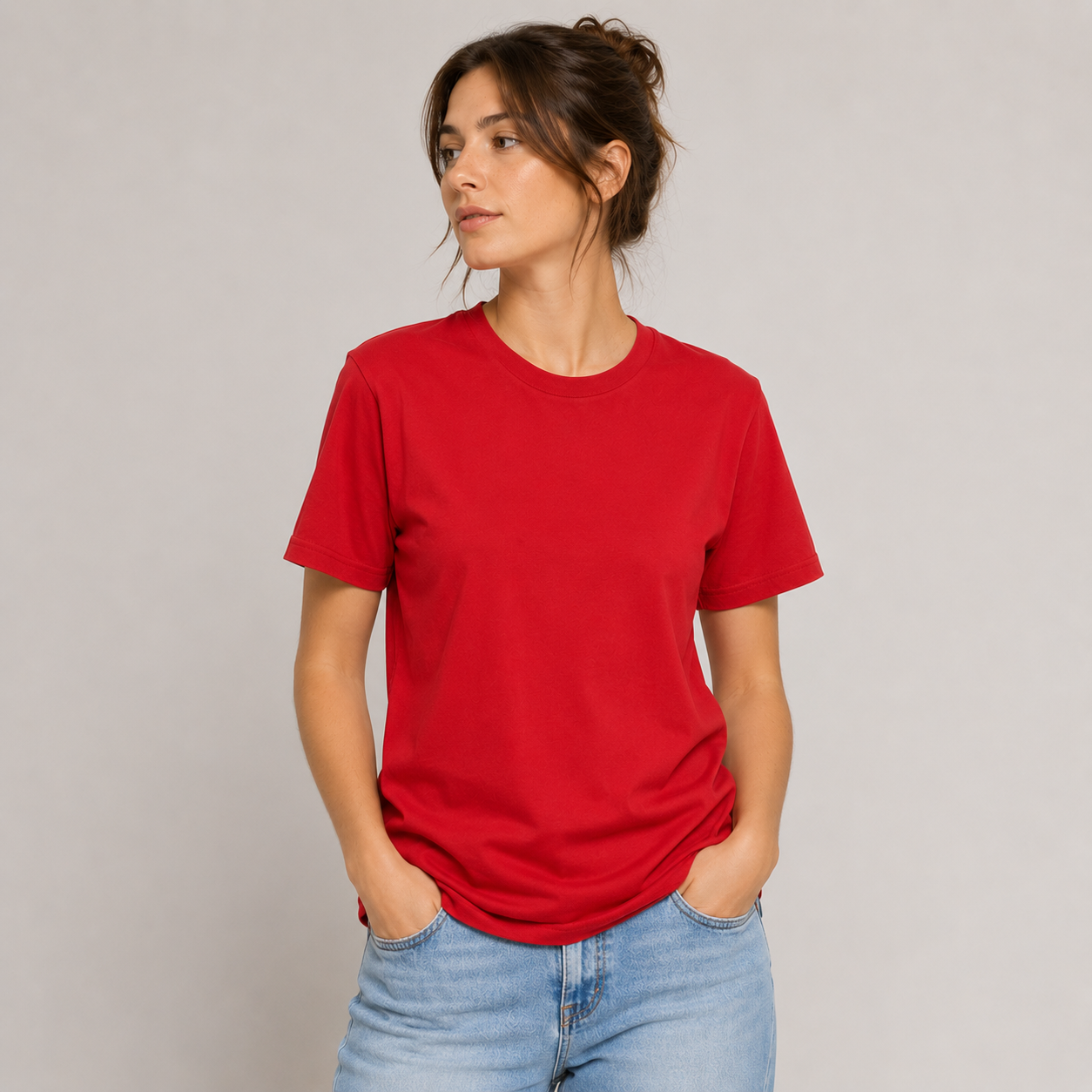 Mujer con camiseta roja y vaqueros de pie con las manos en los bolsillos sobre un fondo gris liso.