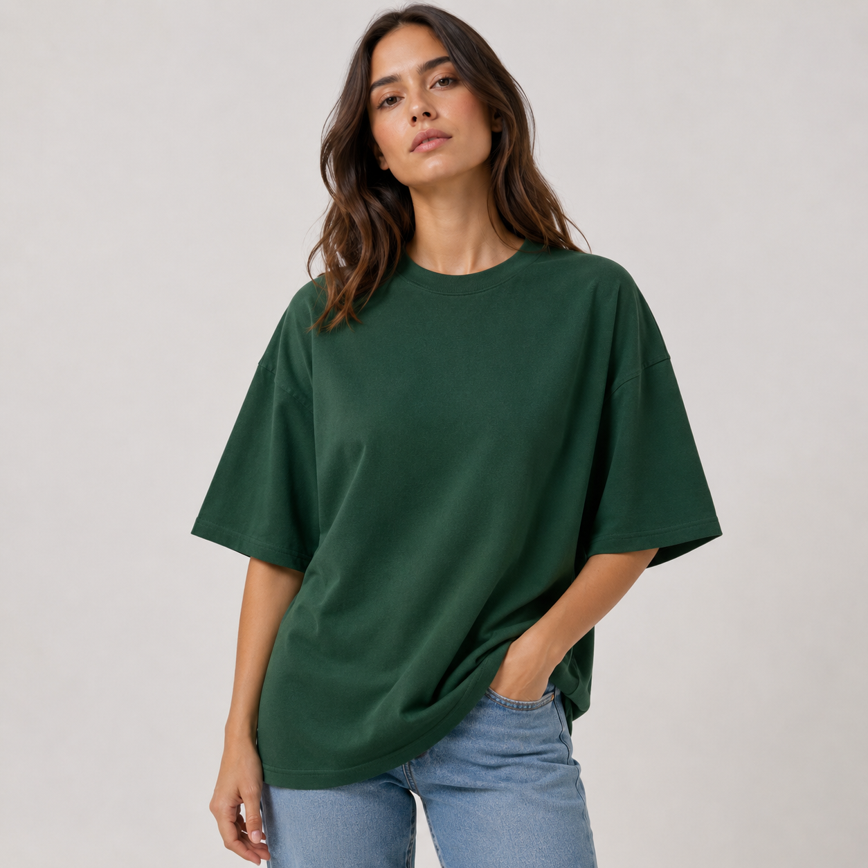 Mujer con una camiseta extragrande de color verde oscuro y vaqueros, de pie sobre un fondo claro liso.