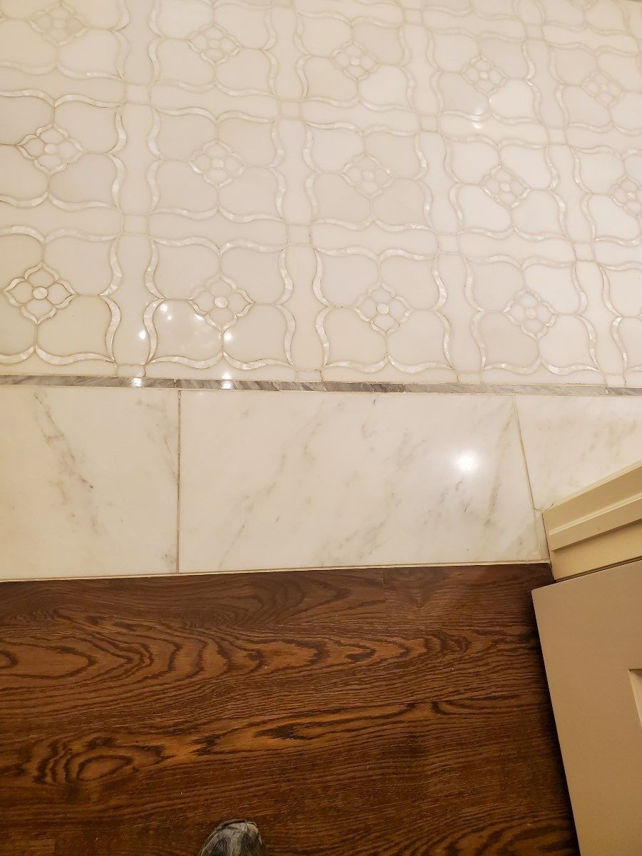 White tiled wall above a beige marble baseboard and dark wood floor beside a cardboard box edge
