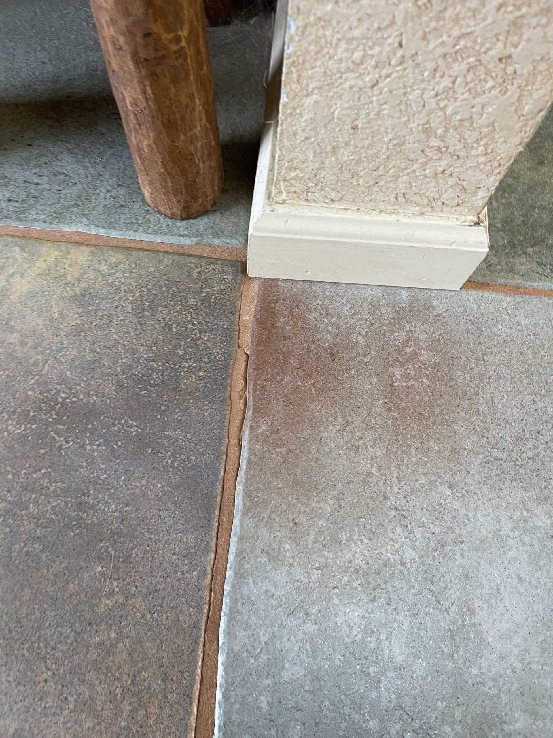 Close-up of a tiled floor with a brown chair leg and a beige wall baseboard corner.
