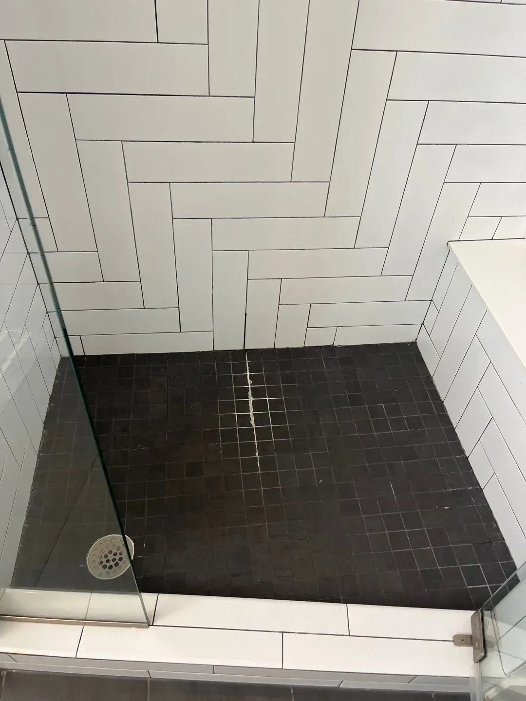 Tiled shower stall with white herringbone walls, black floor tile, and a glass door panel