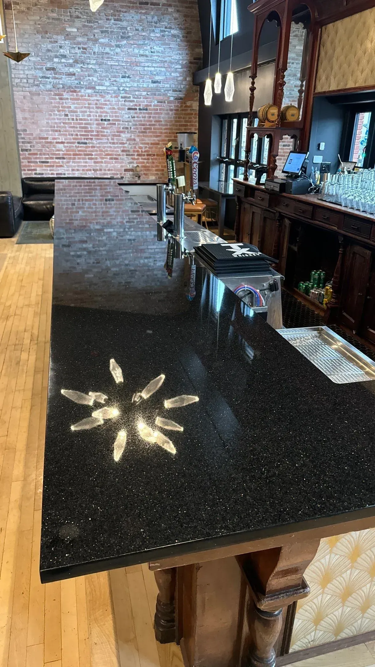 Empty bar counter with black speckled surface and sunlight casting a star-shaped pattern; brick wall and stools nearby