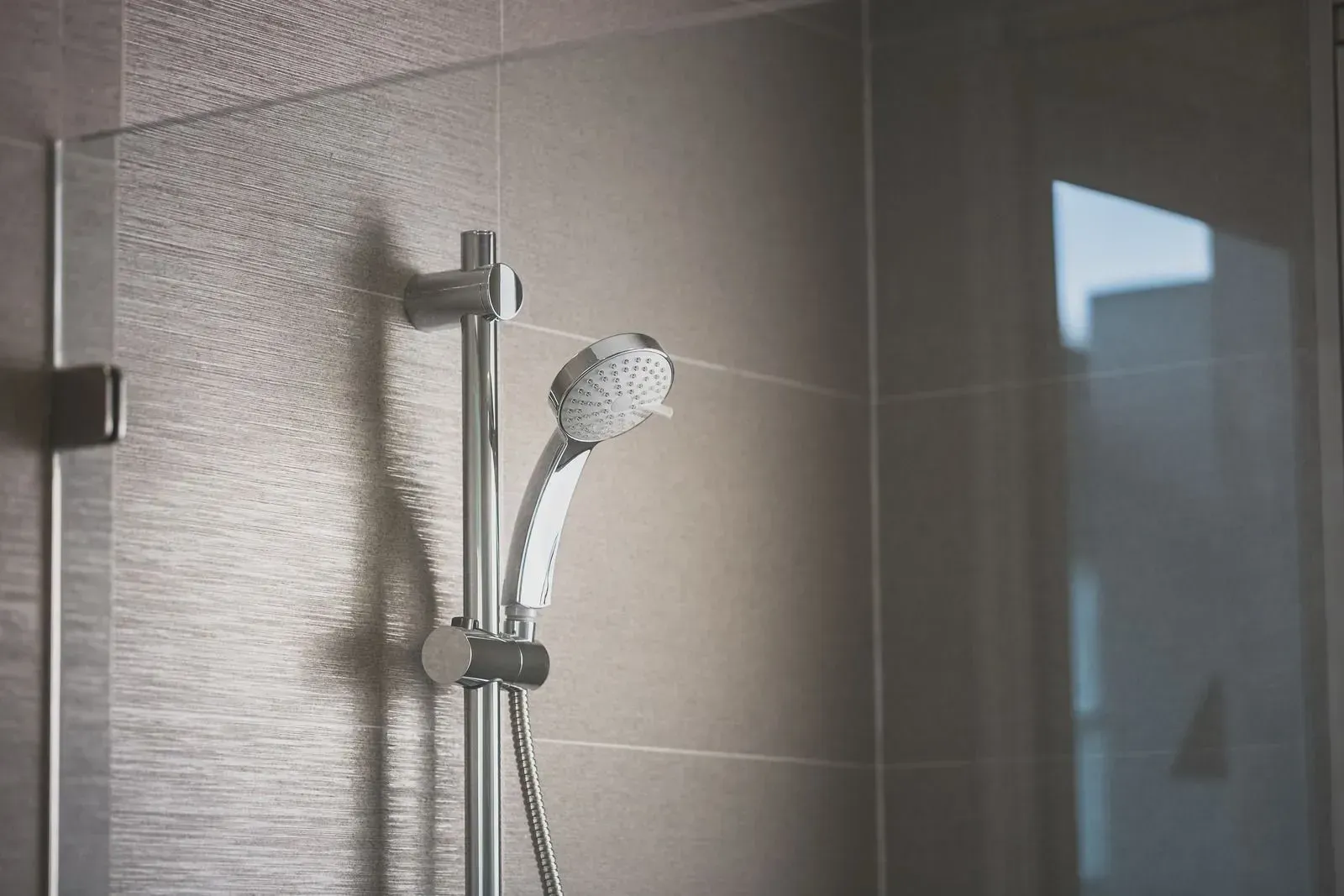 Shower head and hose mounted on a tiled bathroom wall beside a glass shower door.