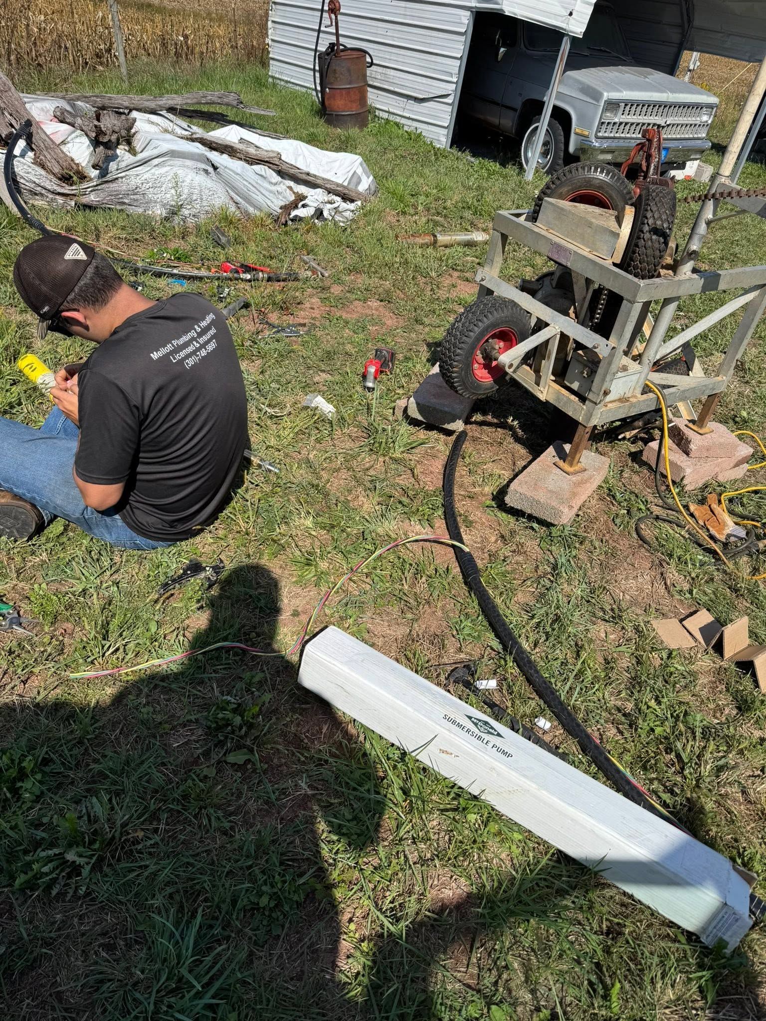Person crouches in a grassy yard beside tools, hoses, and a small machine near a house.