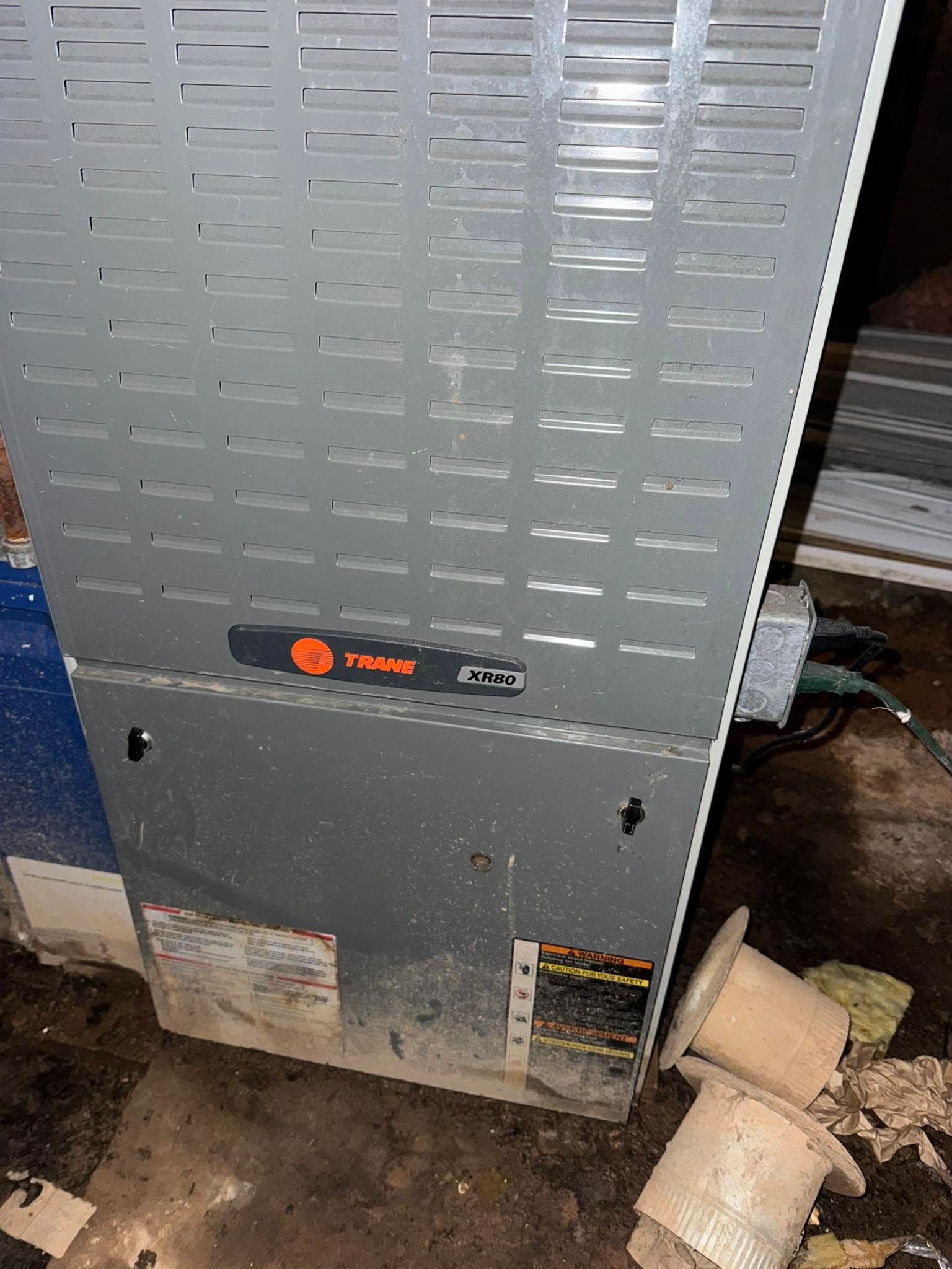 Gray HVAC furnace in a utility area, with visible ductwork and pipes on the floor.