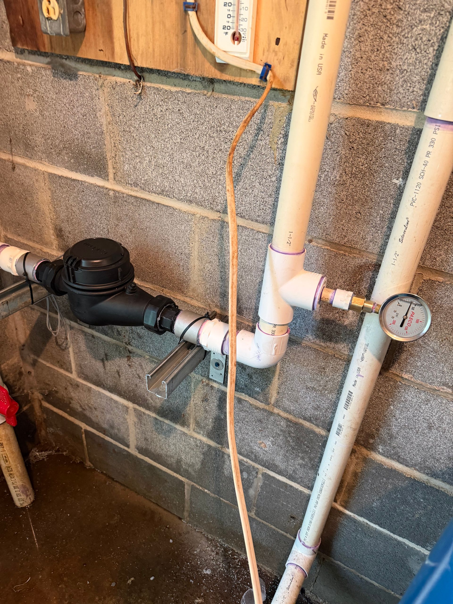 Basement plumbing with white PVC pipes, valve, and pressure gauge against a cinder block wall