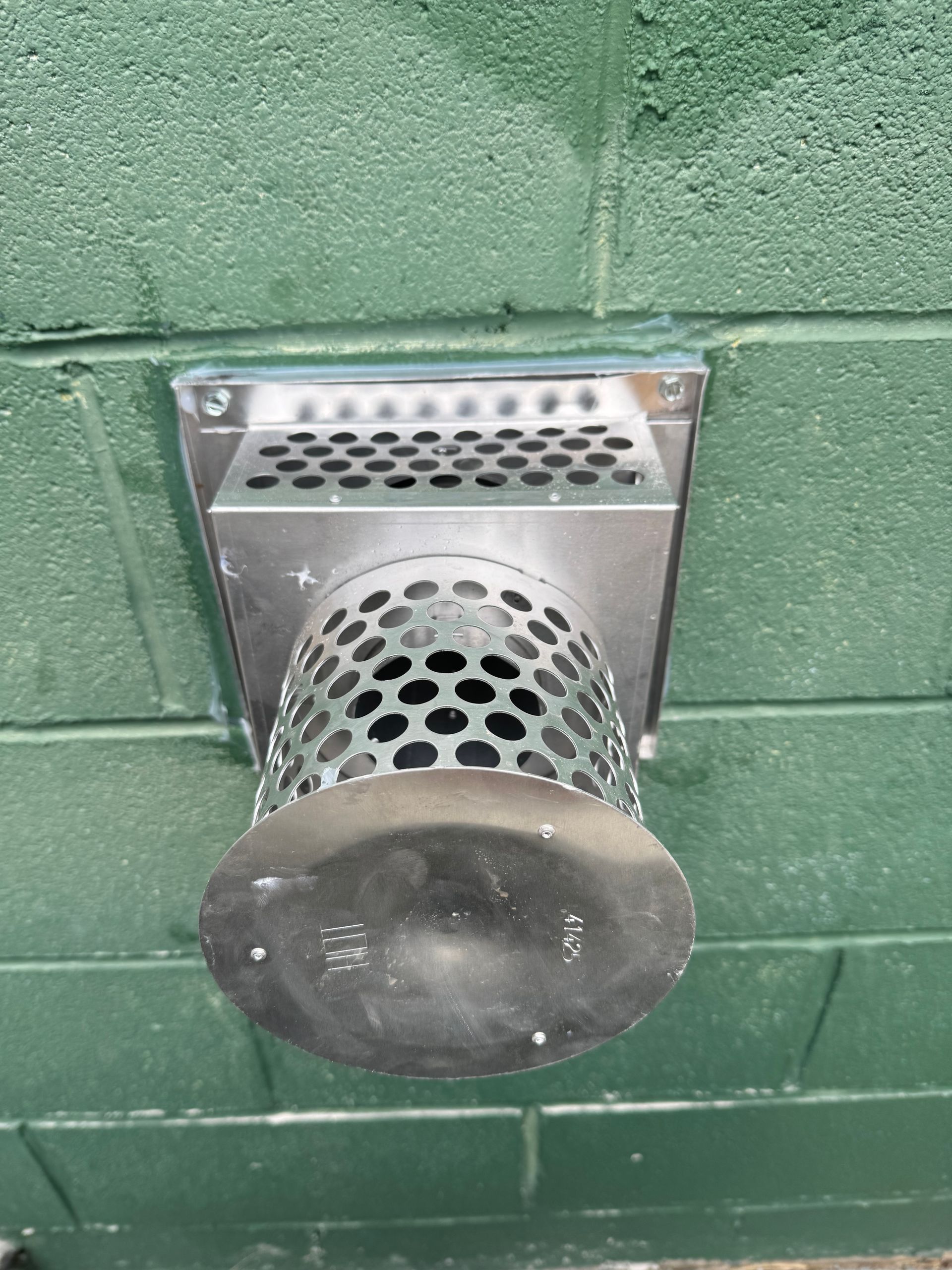 Metal wall vent with a round perforated outlet mounted on a green painted cinder block wall
