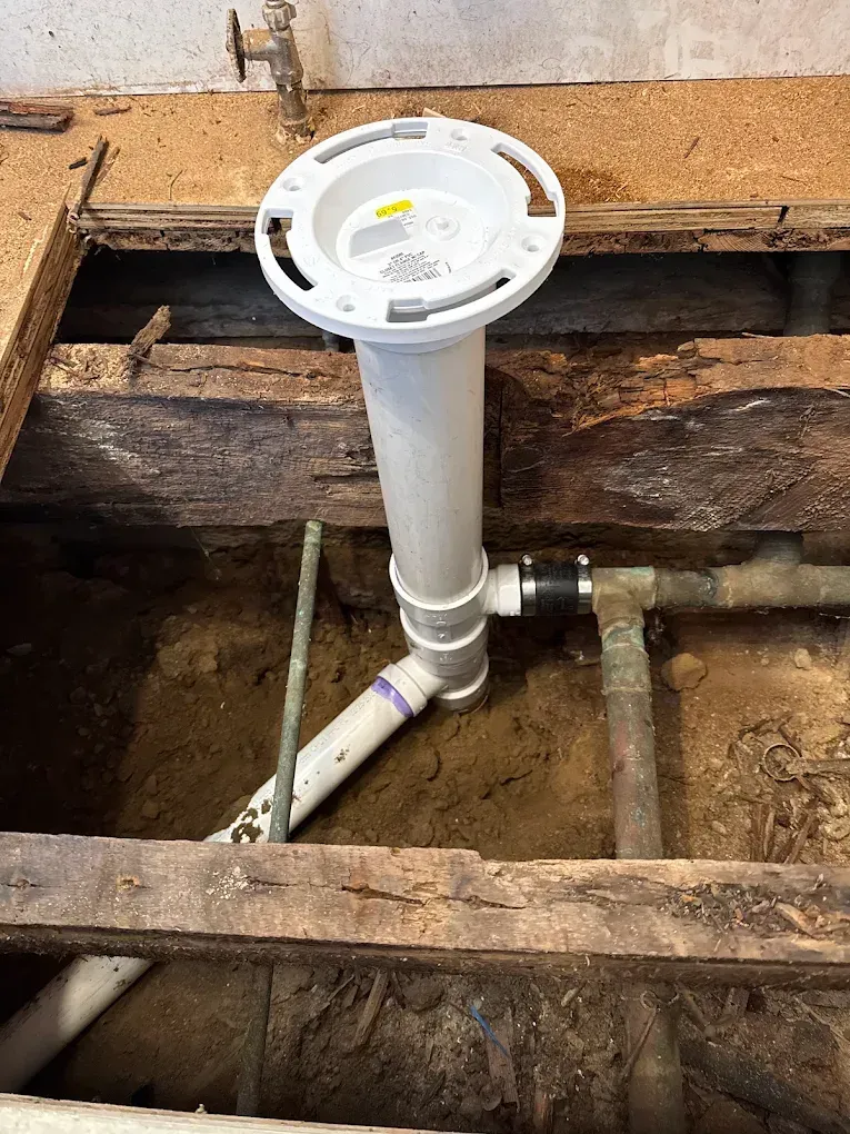 White PVC plumbing vent pipe installed in a floor opening with exposed wood framing and drain pipes