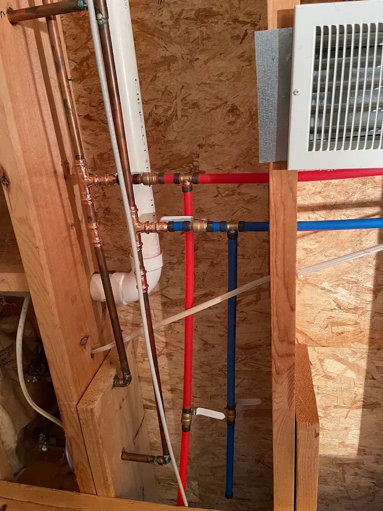 Utility pipes and red-blue PEX lines installed in a wall cavity beside a vent grille.