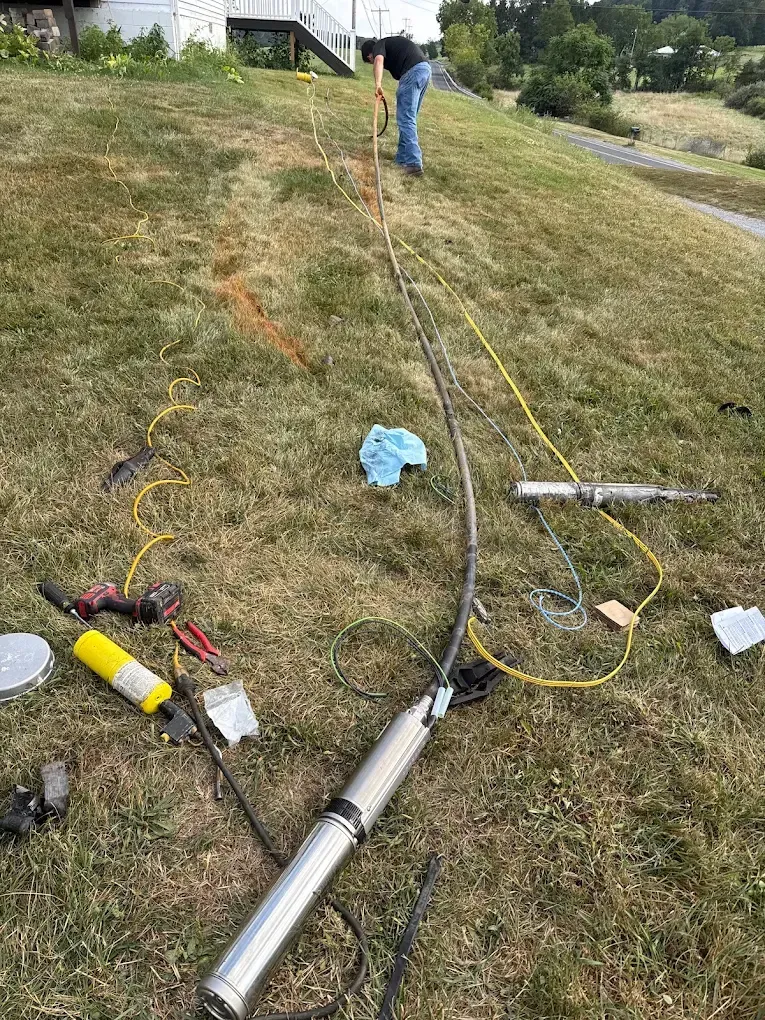 Worker on grassy roadside handling cables and tools near a long metal pipe.
