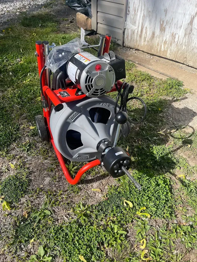 Red engine-driven machine with a large reel sits on grass beside a concrete wall.