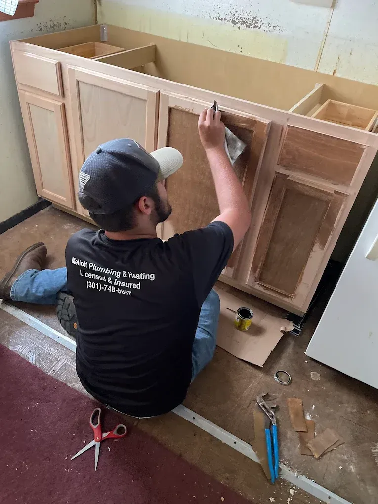 Person installing a light wood cabinet base in a workshop, using a hand tool while kneeling on the floor.