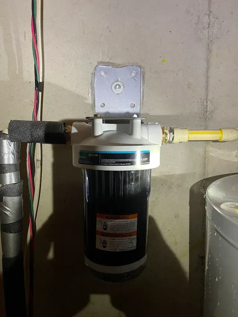 Wall-mounted water filtration unit with black canister, gray fittings, and yellow pipes in a utility room