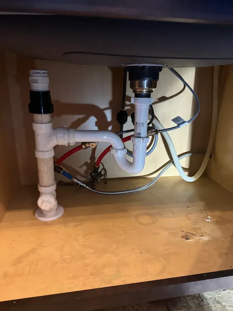 Under-sink plumbing with white PVC pipes, drain trap, and water supply lines in a cabinet.