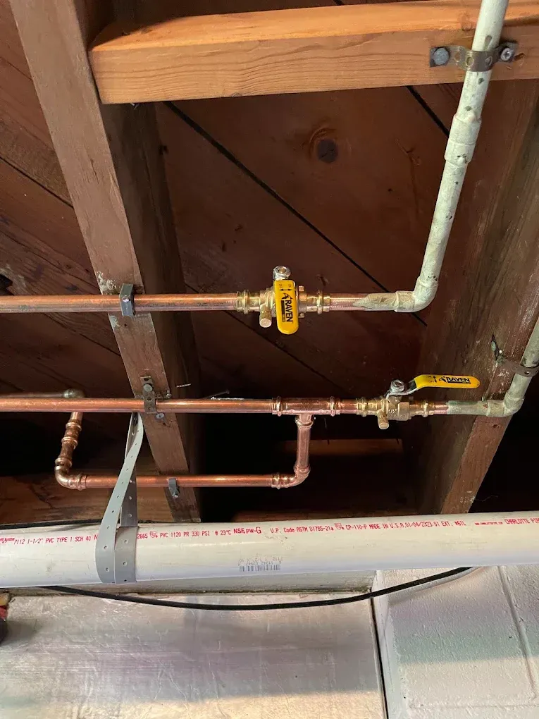 Exposed copper pipes and yellow shutoff valves beneath wooden floor joists in a basement ceiling