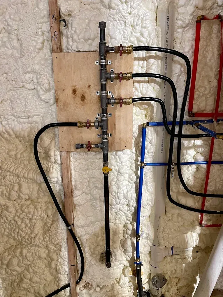 Exposed plumbing and water pipes mounted on a basement wall with insulation and wooden backing.