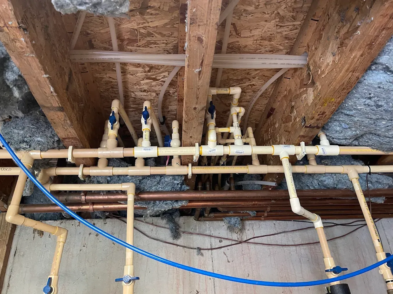 Exposed plumbing pipes and valves beneath a wooden floor, with blue and beige PEX lines.