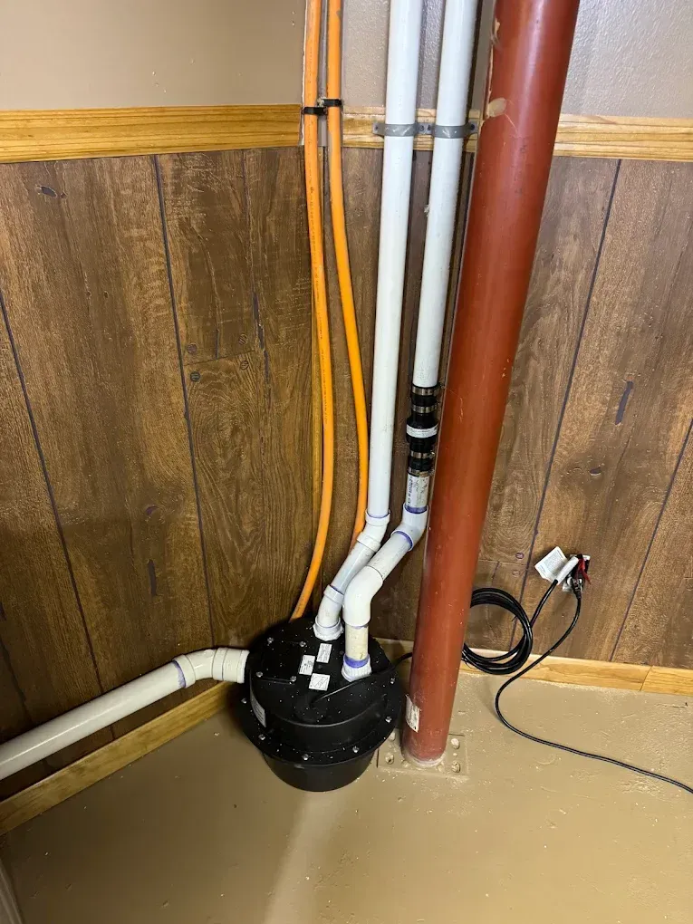 Basement corner with exposed pipes and wires beside a black sump pump.