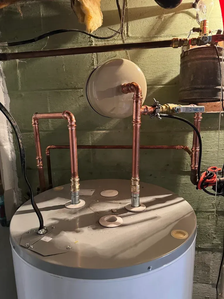 Water heater with copper pipes and a round vent in a basement utility area.