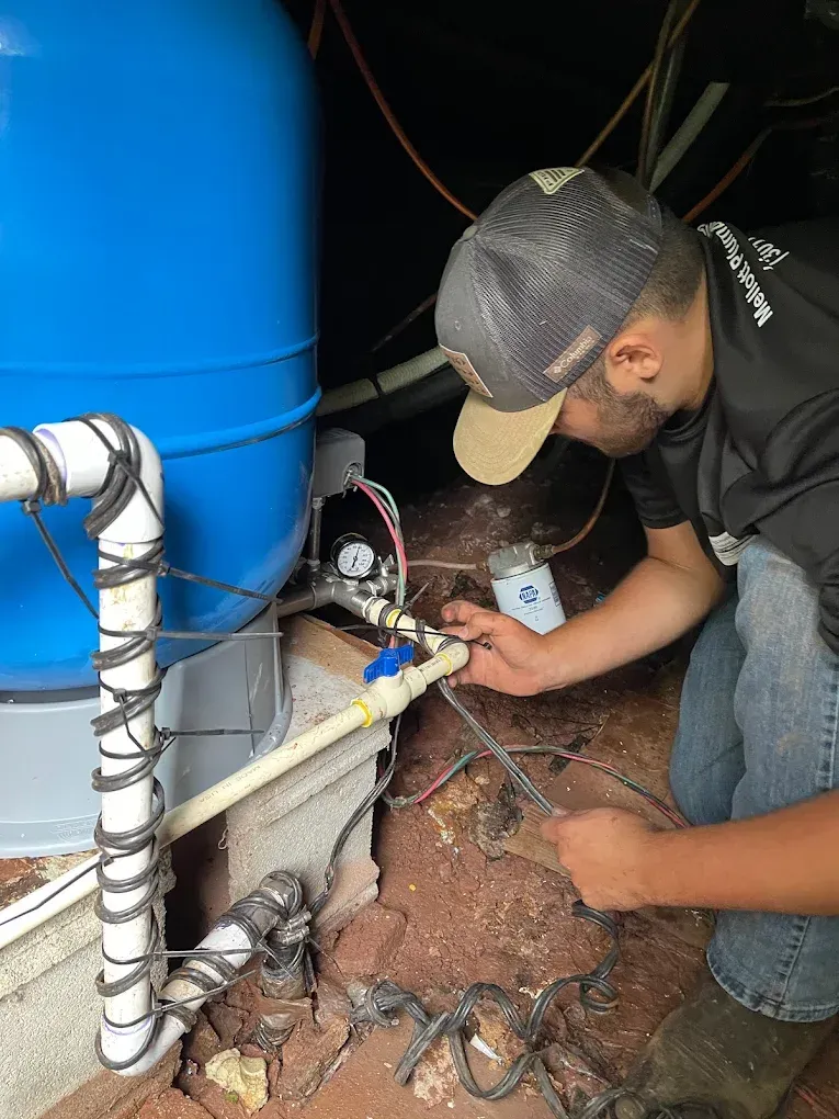 Worker crouches beside blue tank, connecting pipes and wiring near a grounded utility setup