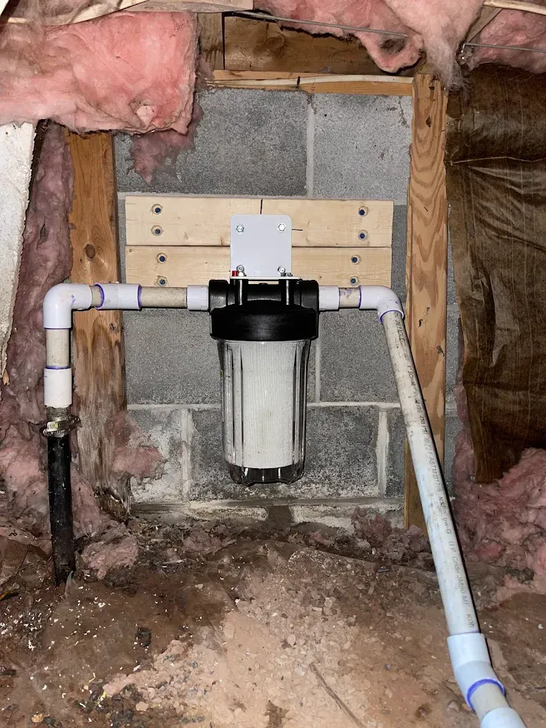 Water filter with white PVC pipes mounted on a cinder block wall in an unfinished basement, under pink insulation.