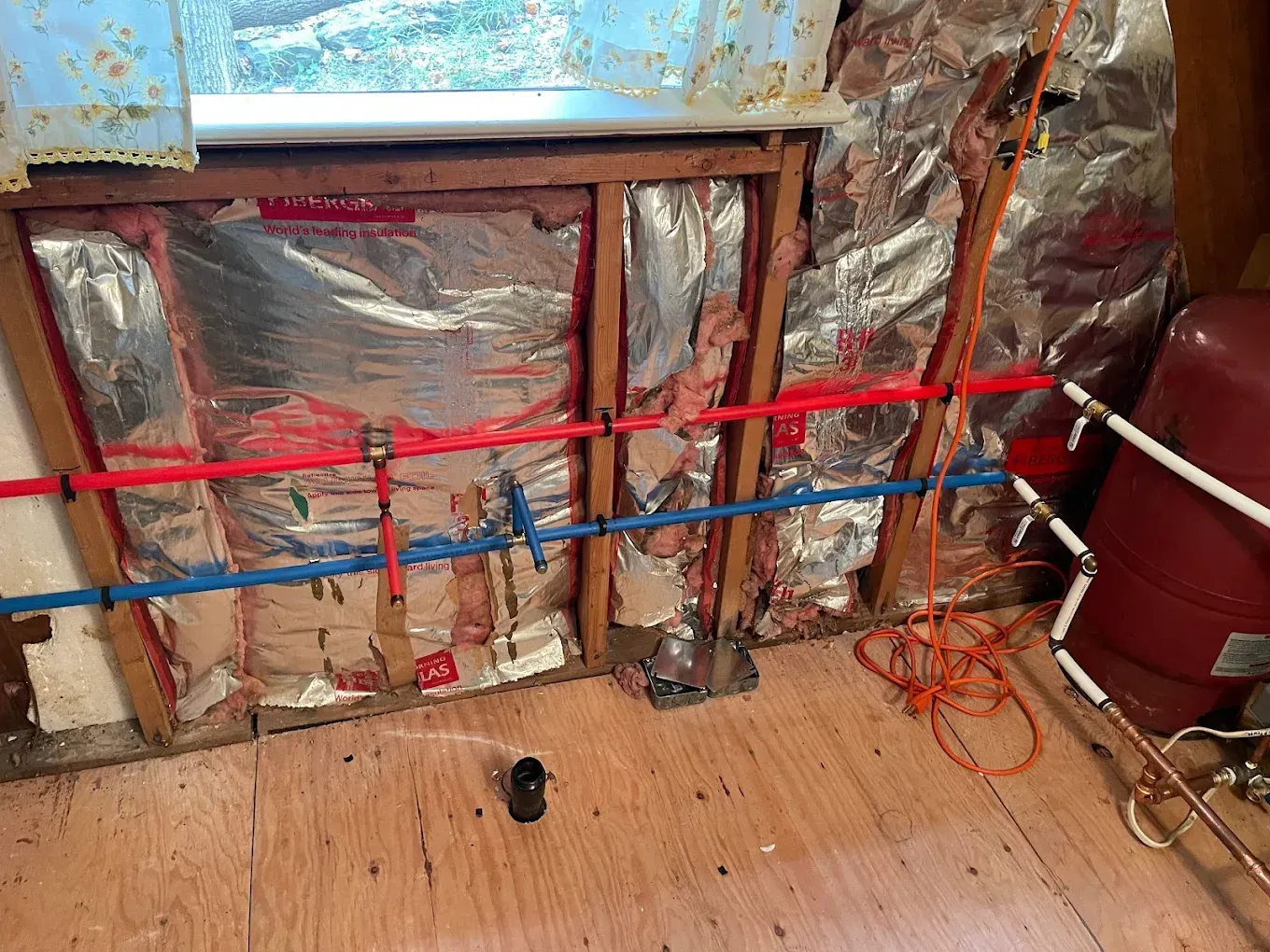 Unfinished wall with insulation, red and blue piping, and a bare plywood floor in a renovation space