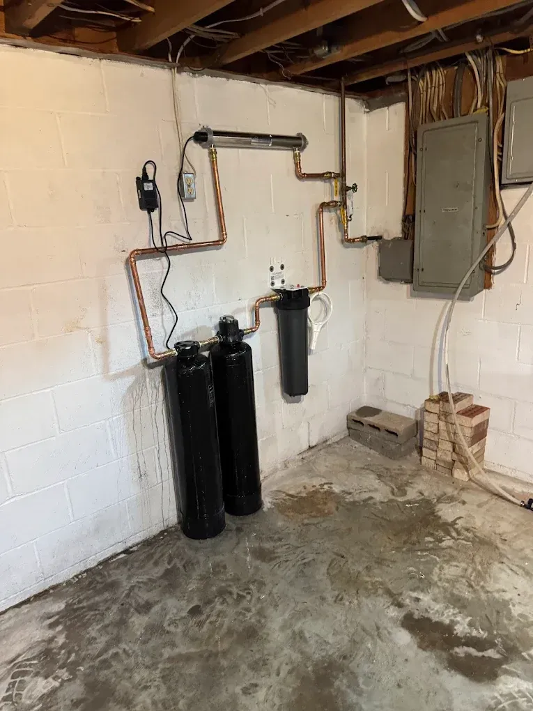 Basement utility wall with pipes, water filters, and electrical panel over a concrete floor.